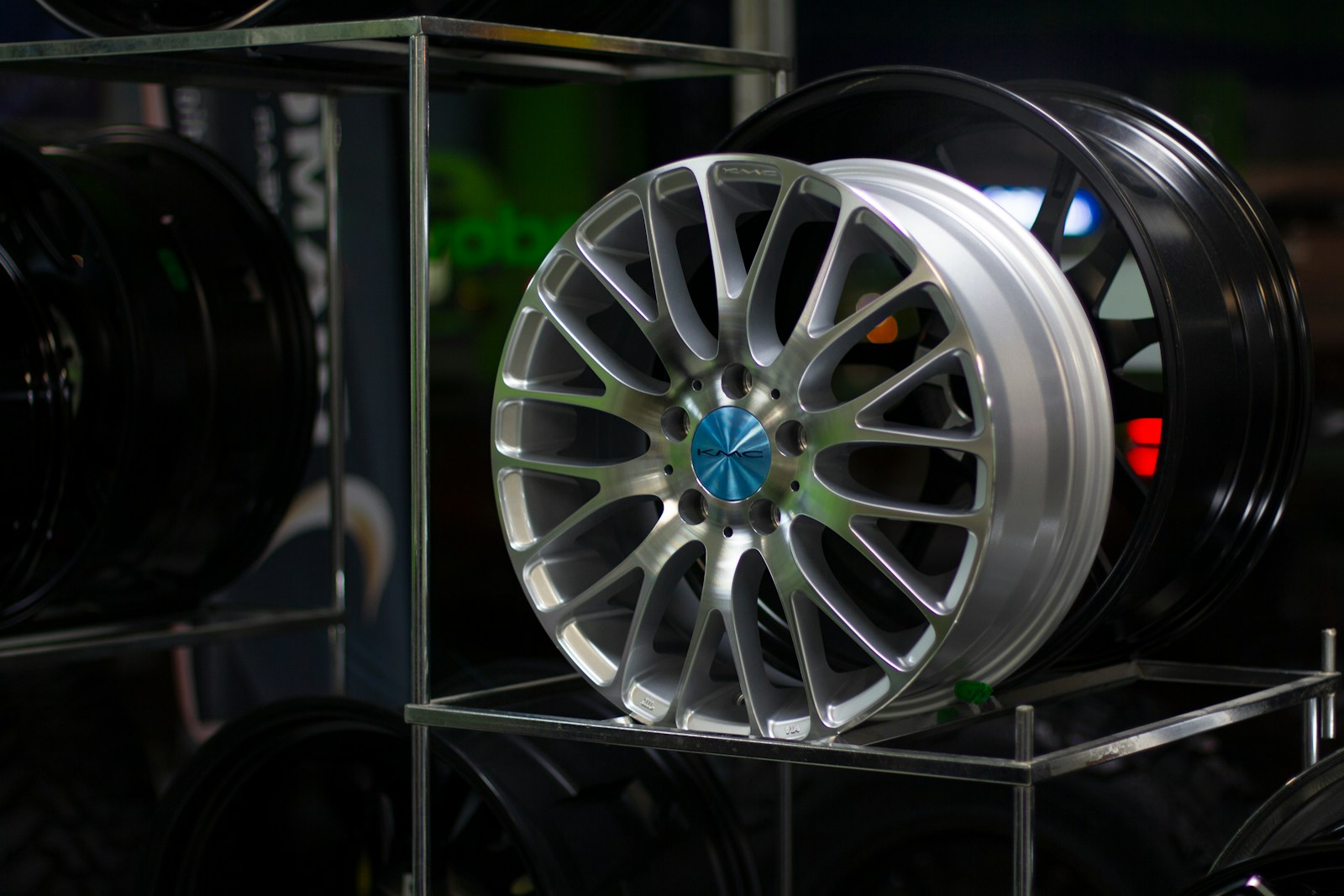 Shiny silver alloy wheel with a blue center cap displayed on a rack alongside black wheels, highlighting automotive customization options.