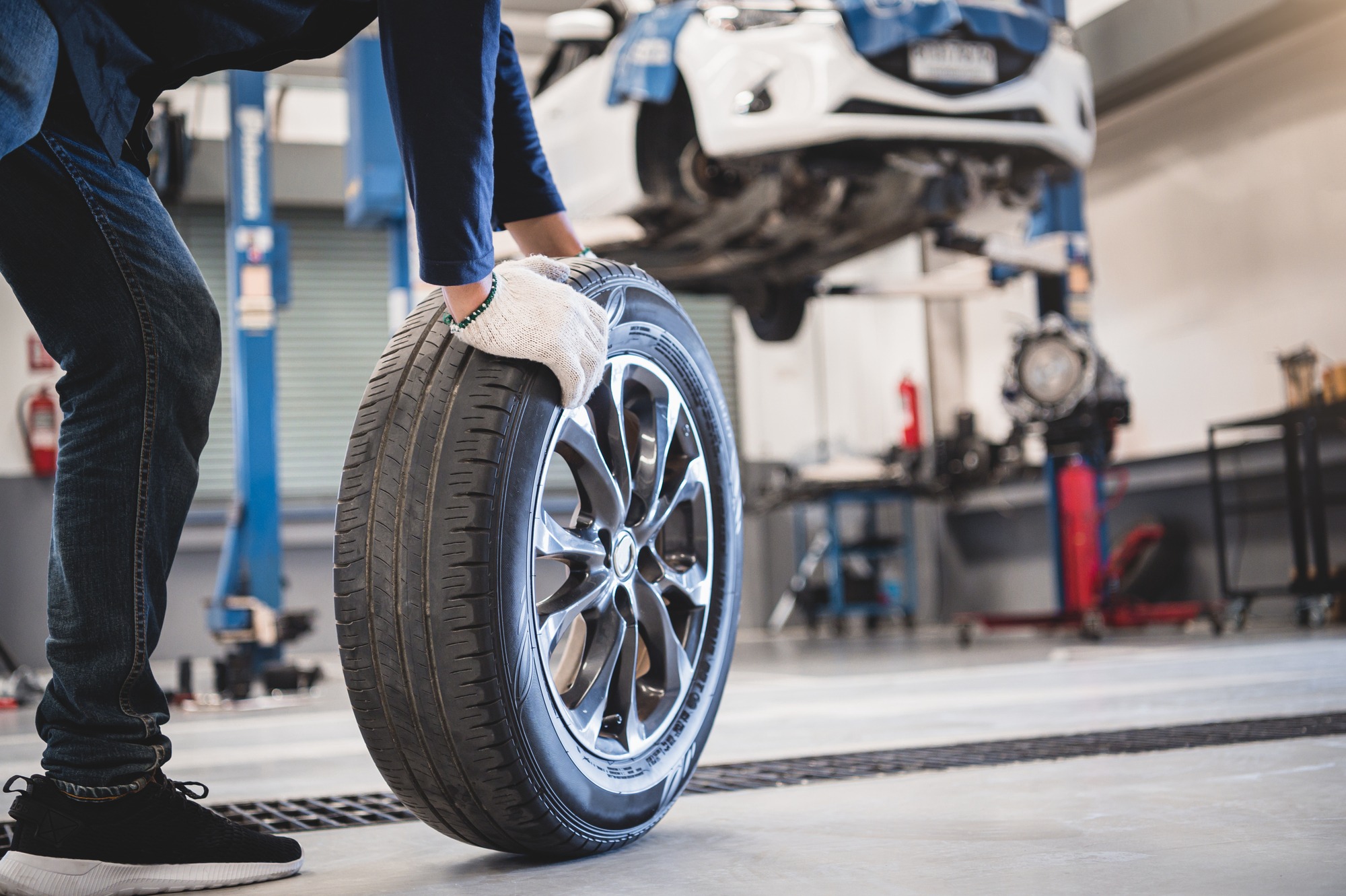 A mechanic wearing gloves lifts a car tire in a workshop, with a lifted car in the background, showcasing tire change or maintenance activity.