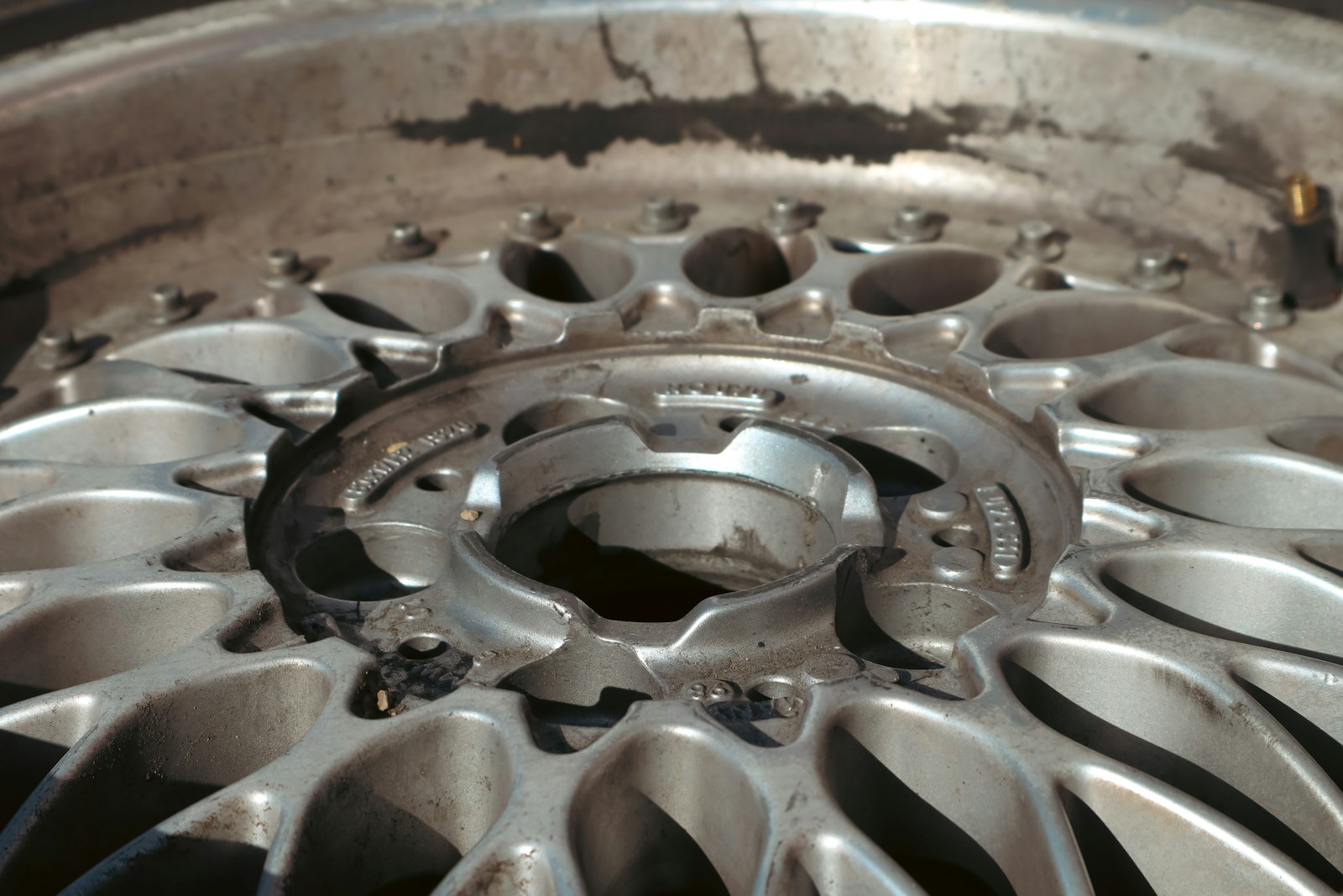 Close-up of a dirty, metallic wheel rim with intricate spoke design, showing signs of wear and age. Relevant for automotive discussions or repairs.